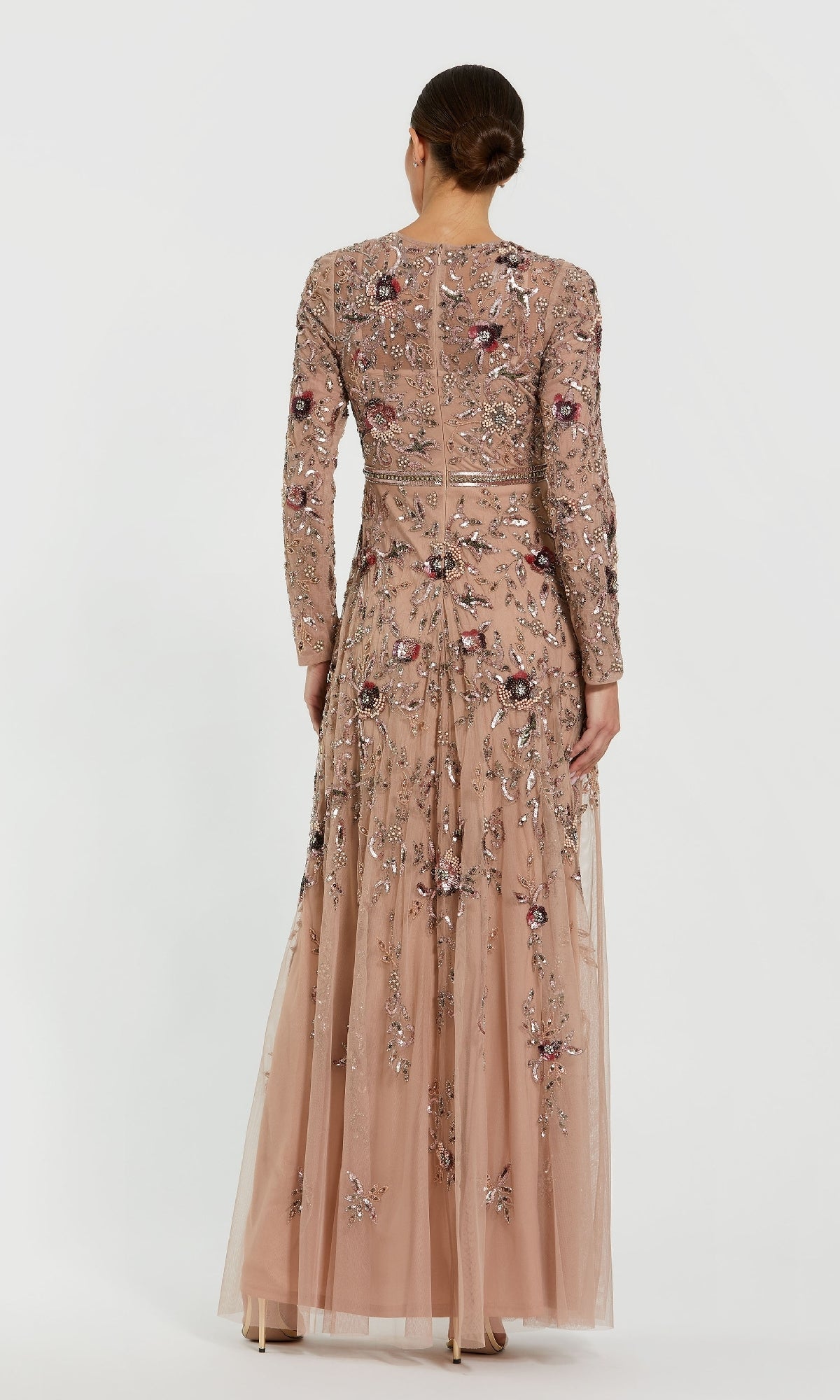 A woman stands with her back to the camera, wearing the Mac Duggal 10093 Long Sleeve Beaded Formal Dress in a sheer nude shade with intricate floral embroidery and beading, her hair styled neatly in a bun.